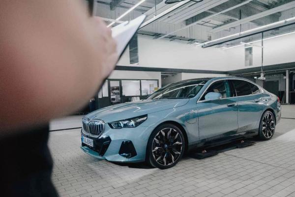 Ein hellblauer BMW steht auf der Hebebühne in einer BMW-Werkstatt. Ein Mitarbeiter füllt den Regiezettel aus.